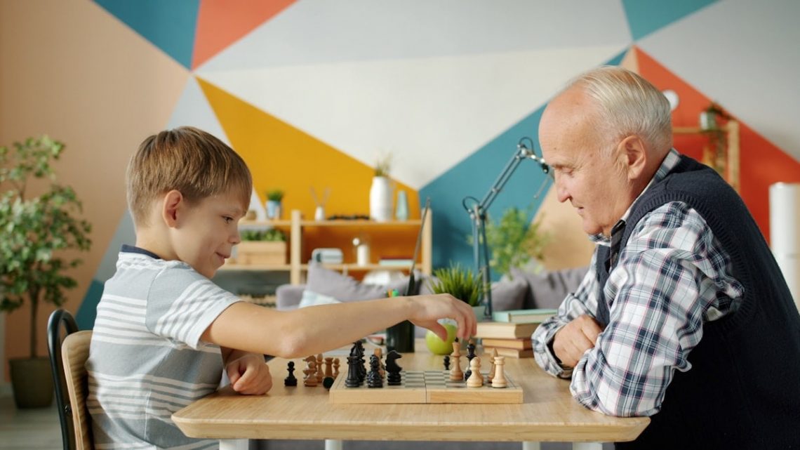 Cheerful people old man and adorable child are playing chess in apartment talking smiling enjoying game. Entertainment, leisure time and family concept.