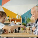 Cheerful people old man and adorable child are playing chess in apartment talking smiling enjoying game. Entertainment, leisure time and family concept.