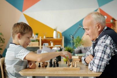 Cheerful people old man and adorable child are playing chess in apartment talking smiling enjoying game. Entertainment, leisure time and family concept.