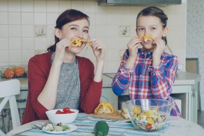 Joyful mother and cute cheerful daughter have fun grimacing silly with vegetables and looking into camera while cooking in the kitchen at home. Family, cook, and people concept